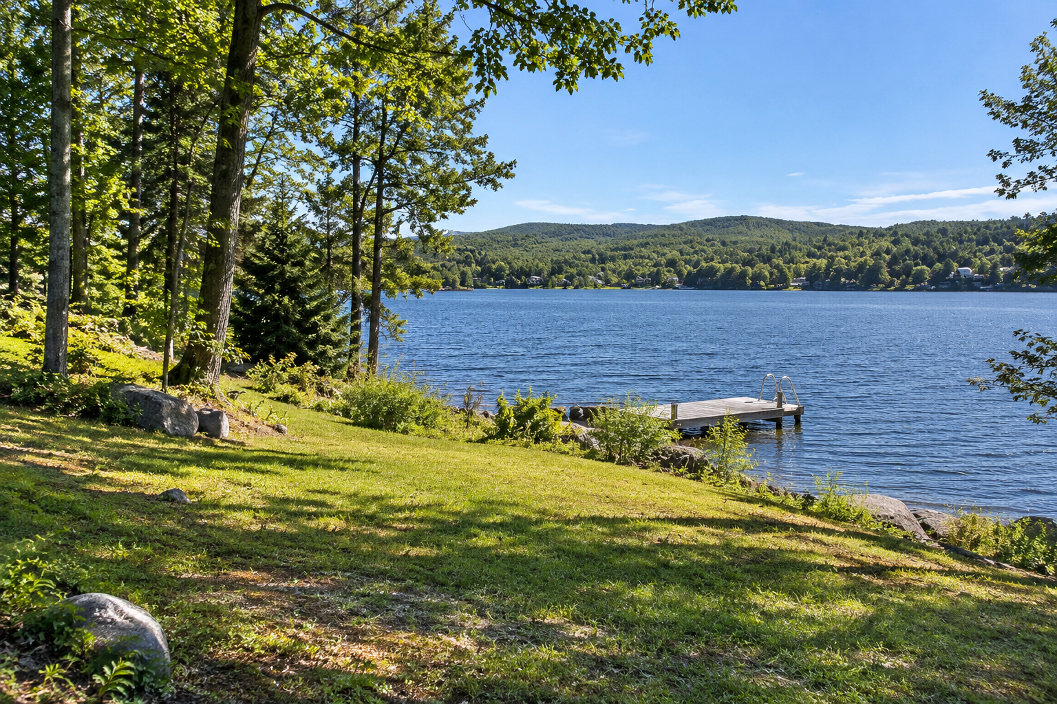 Terrain gazonné et arboré en pente douce au bord d'un lac, avec petit quai, rive rocheuse et collines boisées.